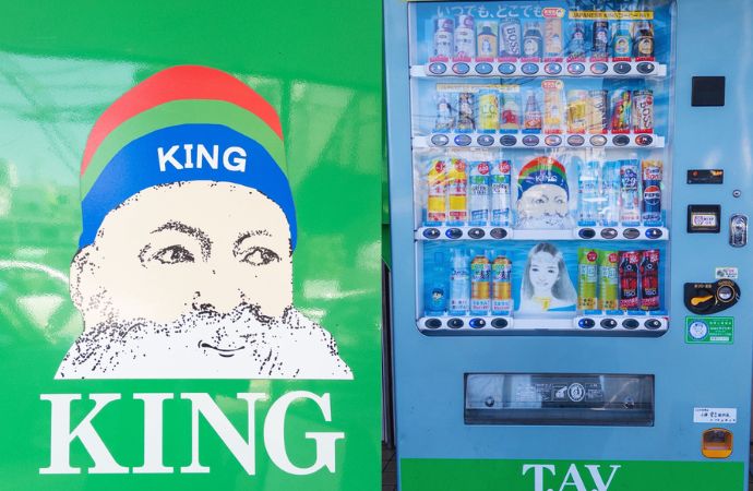 名古屋市内で見られる『KINGコーヒー』の自販機の実物写真。左側には鮮やかな水色の筐体に王冠を被った髭の男性のイラストが描かれたインパクトのあるKINGコーヒー専用機、右側には一般的な飲料メーカーの自販機が並んでいる。自販機を『棚』として活用し、自社ブランドの格安飲料を提供する新興勢力の象徴的な光景。